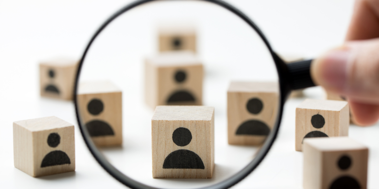 Symbolbild: Teilnehmende gesucht (eine Person hält eine Lupe vor Holzklötze mit Personen)