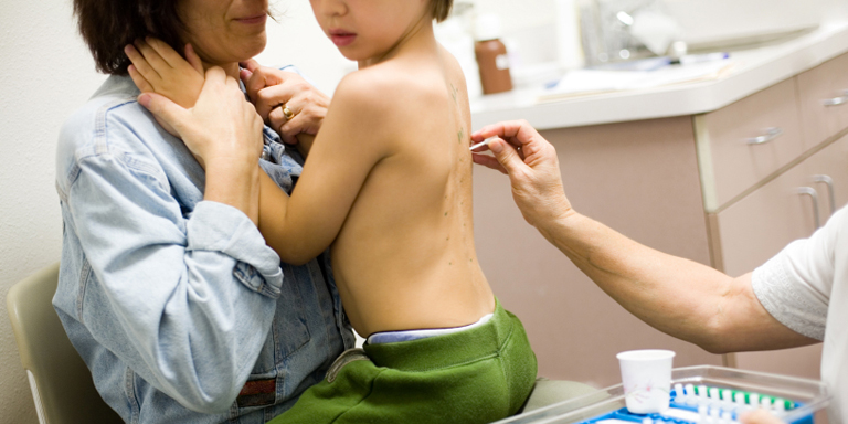 Doctor doing prick tests on a child's back. The child sits on his mother's lap.