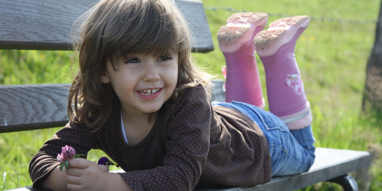 Una bambina è sdraiata su una panchina, con le gambe piegate e guarda contenta il mondo